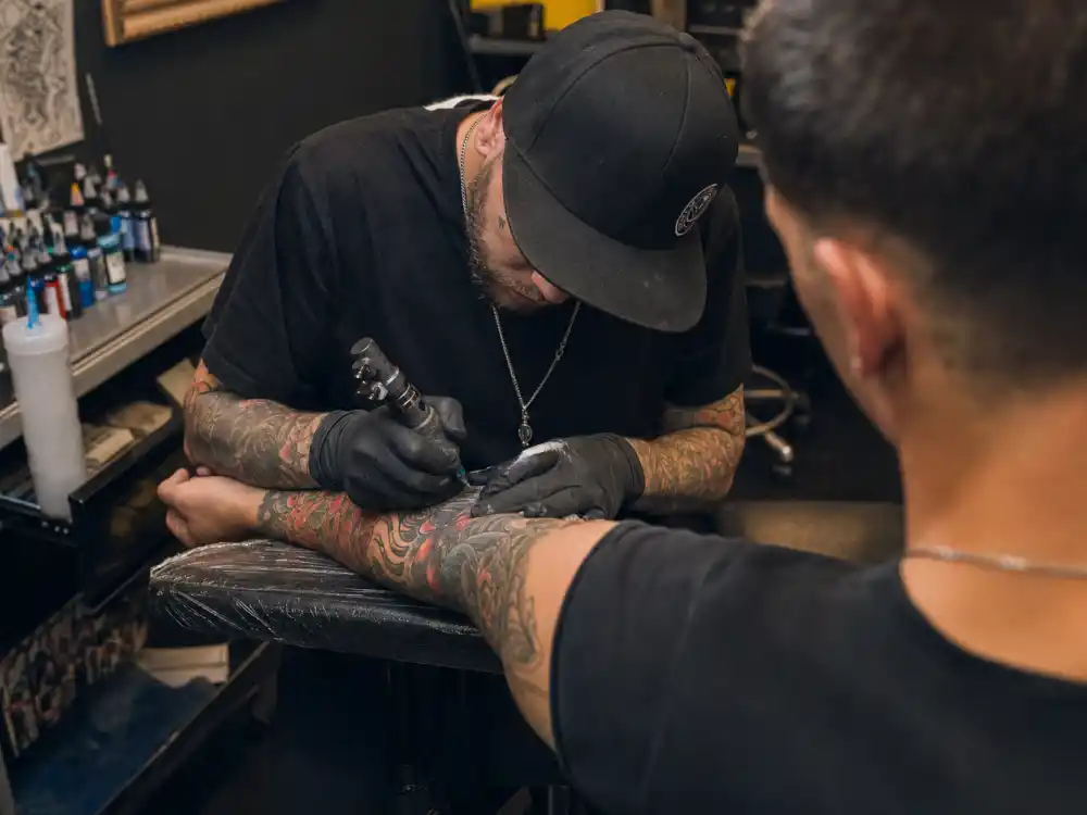 Lionel Messi tattoos and their meanings studio session — tattoo artist gloved hands applying ink to forearm of athletic client over-shoulder angle