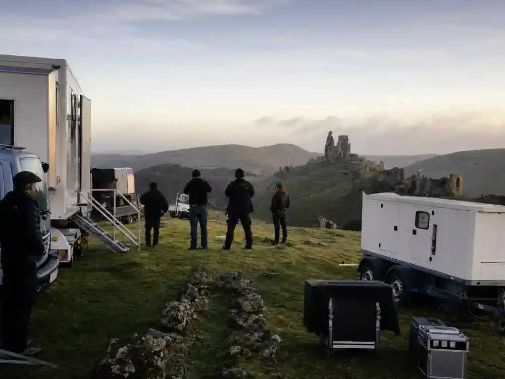Lord of the Rings Rings of Power cast guide filming locations — UK countryside production trucks and crew silhouettes scouting green hills and ancient stone ruins