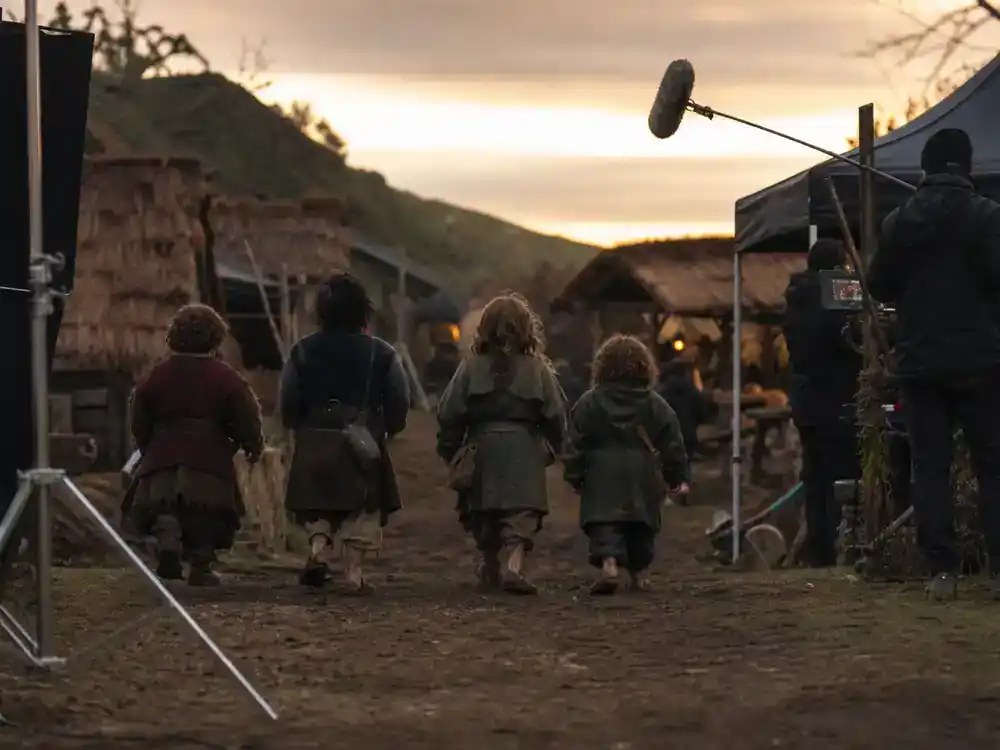 Lord of the Rings Rings of Power cast guide Harfoots ensemble — small cast silhouettes from behind on outdoor fantasy village film set with production tents at dusk