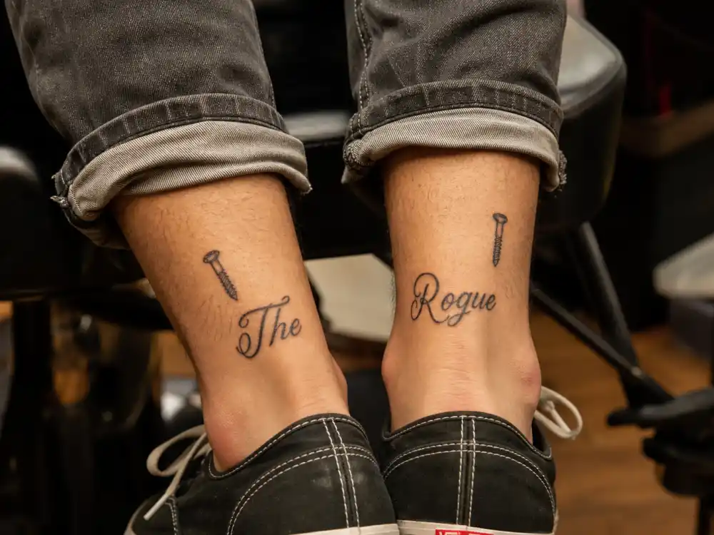 Louis Tomlinson tattoos and their meanings ankle matching — screw tattoos and The Rogue script split across both ankles close-up