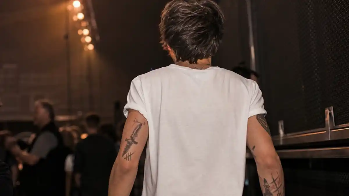 Louis Tomlinson tattoos and their meanings — British singer from behind backstage showing patchwork arm tattoos including dagger and compass