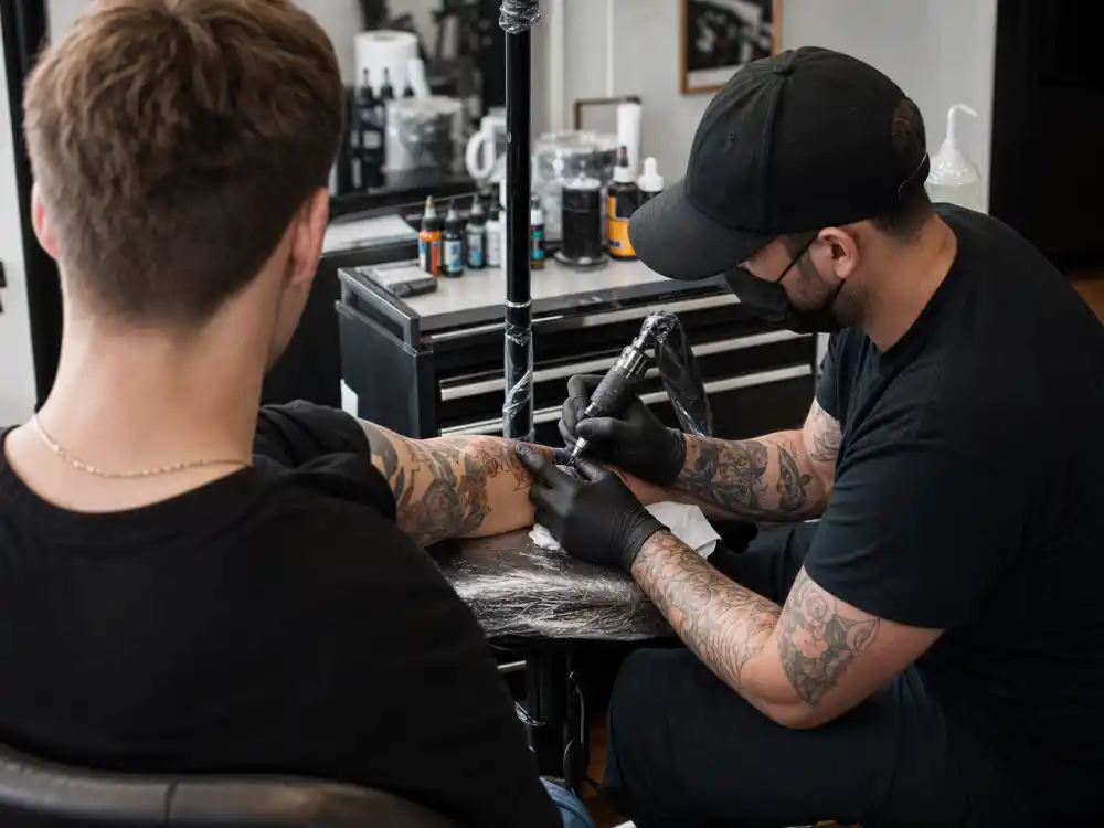Louis Tomlinson tattoos and their meanings studio session — tattoo artist gloved hands applying ink to forearm over-shoulder client angle