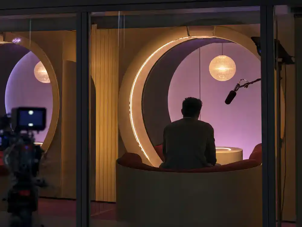 Love Is Blind season 8 cast guide pods — dating show pod room interior with warm colored booths participant silhouette and boom mic visible from behind glass