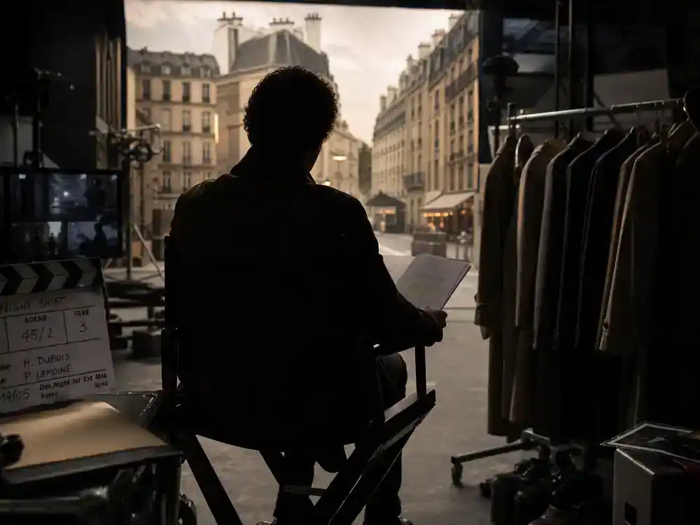 Lupin Netflix cast and episode guide Assane Diop — lead actor silhouette from behind on director chair reviewing script on Paris TV heist drama soundstage
