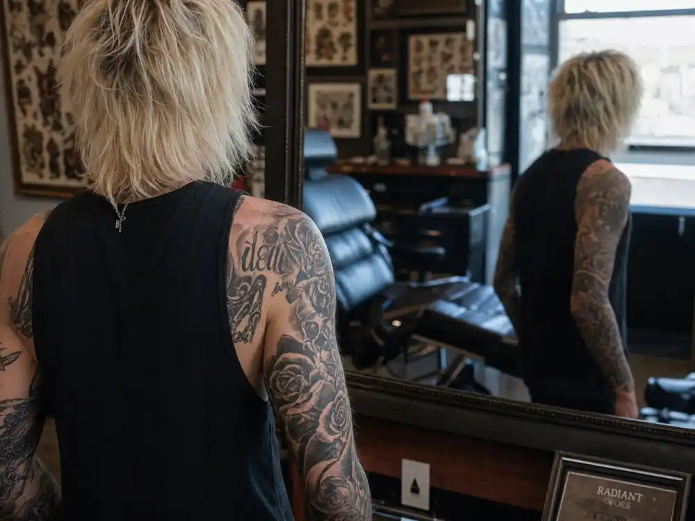 Machine Gun Kelly tattoo collection guide arm sleeve — over-shoulder mirror view of roses skulls and script black-grey forearm tattoos on bleached blonde musician