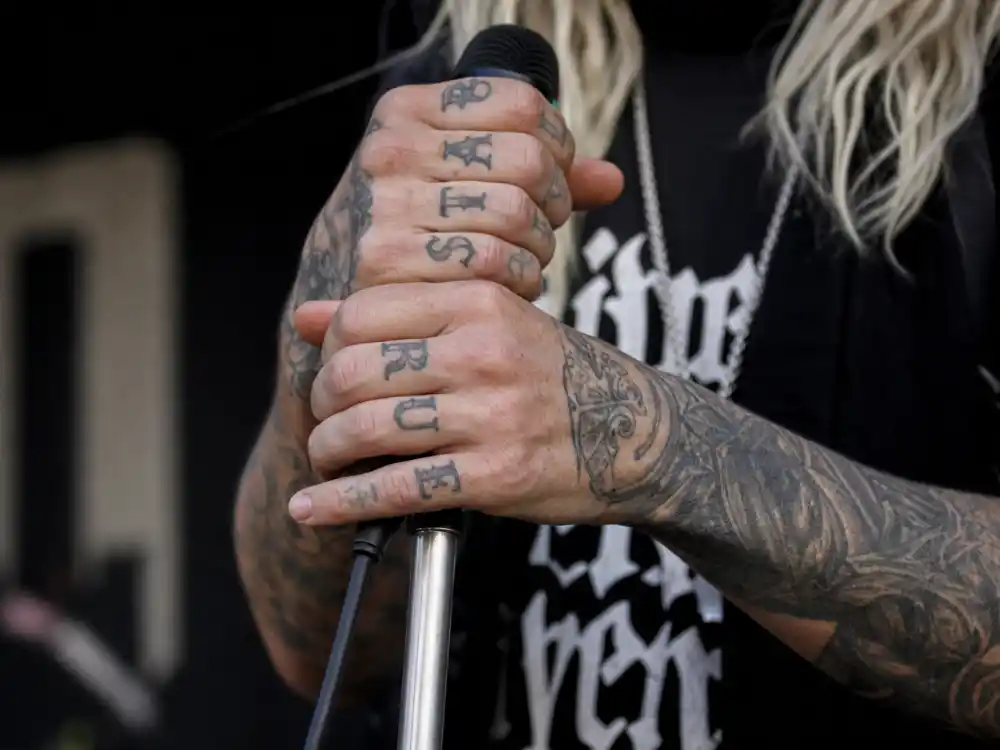 Machine Gun Kelly tattoo collection guide hand ink — close-up of knuckle script and finger lettering on tattooed hands holding microphone with forearm sleeve visible