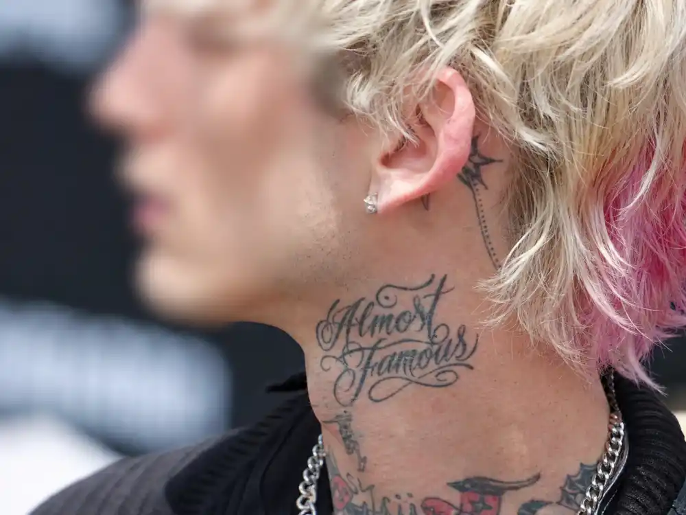 Machine Gun Kelly tattoo collection guide neck ink — blurred side profile showing throat script and behind-ear tattoos on bleached blonde rock musician outdoors