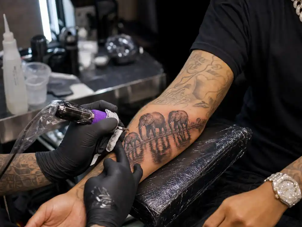 Maluma tattoos and their meanings — four elephants family tattoo with water reflection on right forearm during professional studio session