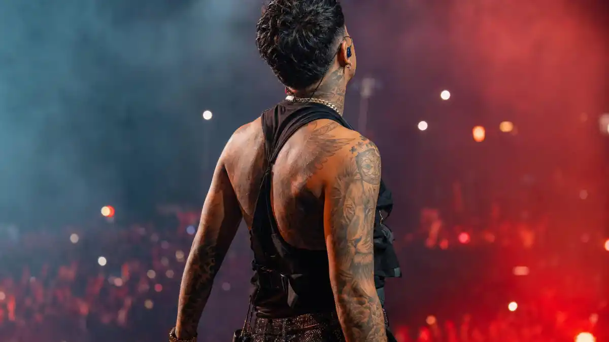 Maluma tattoos and their meanings — Colombian reggaeton star from behind on concert stage with lion chest tattoo and arm sleeves visible under stage lights