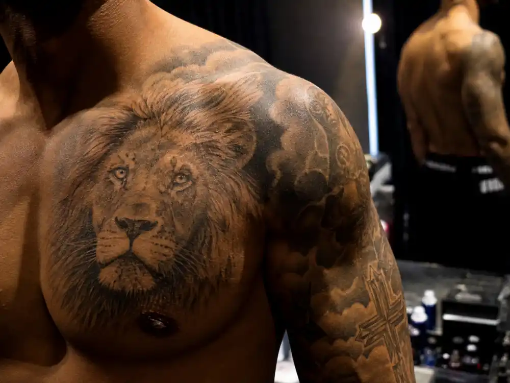 Maluma tattoos and their meanings — large lion tattoo on left chest and shoulder visible in shirtless backstage mirror reflection