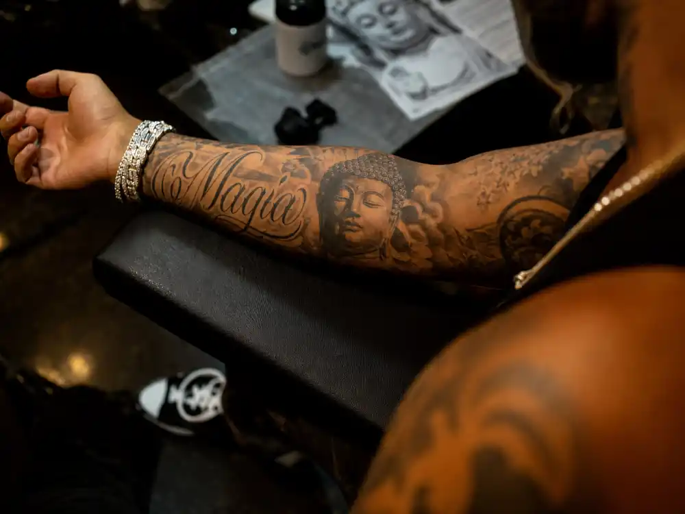 Maluma tattoos and their meanings — close-up of Magia script and Buddha design on left forearm with fine black and grey ink detail