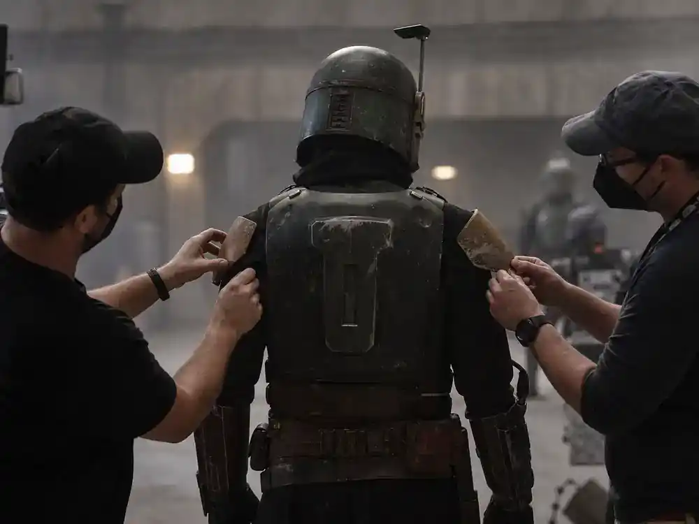 Mandalorian and Grogu movie cast explained Pedro Pascal — actor silhouette from behind in armored bounty hunter costume on film set with crew adjusting helmet