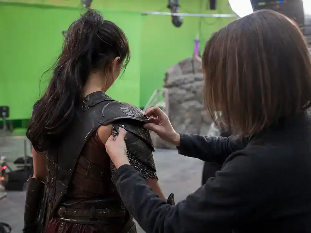 Masters of the Universe movie 2026 cast guide Camila Mendes Teela — warrior costume fitting silhouette from behind on film set with armor department crew