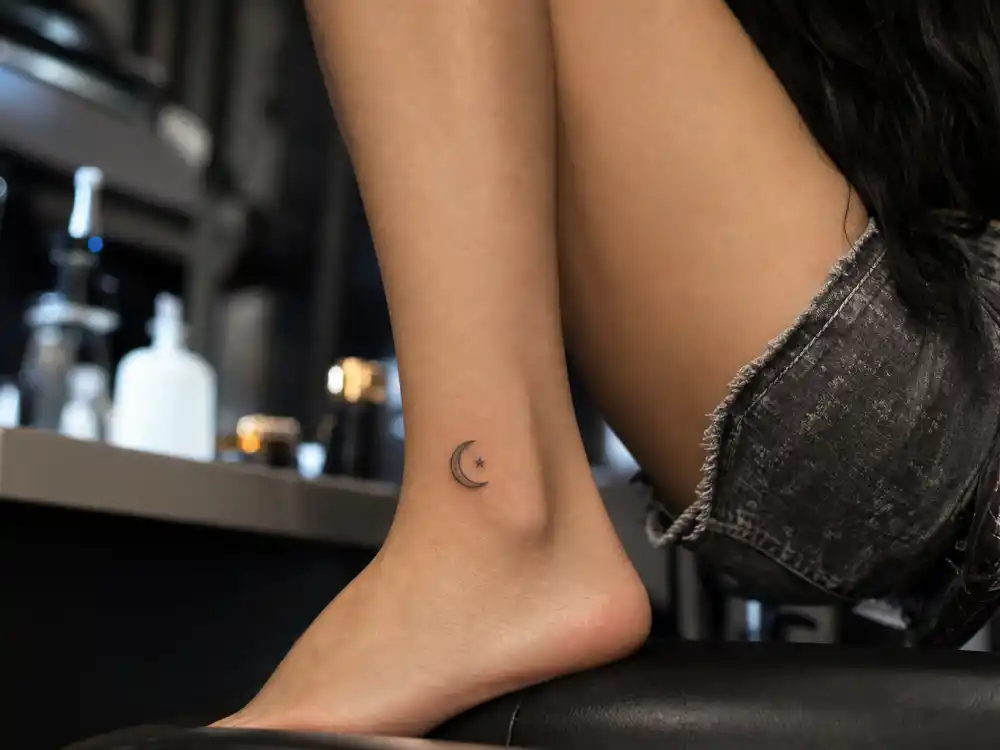 Megan Fox tattoos and their meanings — small moon and star ankle tattoo visible in studio close-up from behind