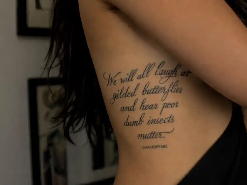 Megan Fox tattoos and their meanings — Shakespeare King Lear script tattoo along ribcage photographed from over-shoulder angle