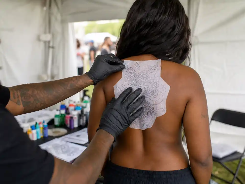 Megan Thee Stallion tattoos and their meanings studio — makeup artist applying temporary transfer body art to woman with long dark wavy hair backstage