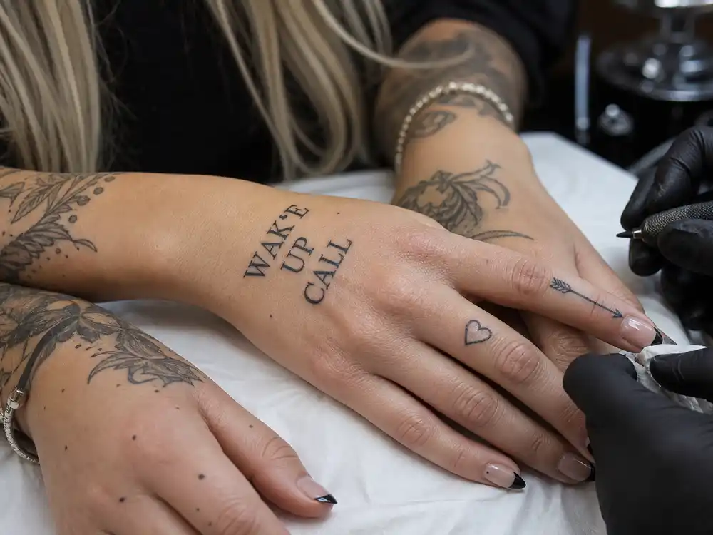Miley Cyrus tattoos and their meanings — close-up of heart finger tattoo, arrow index ink, and WAKE UP CALL hand script in studio session
