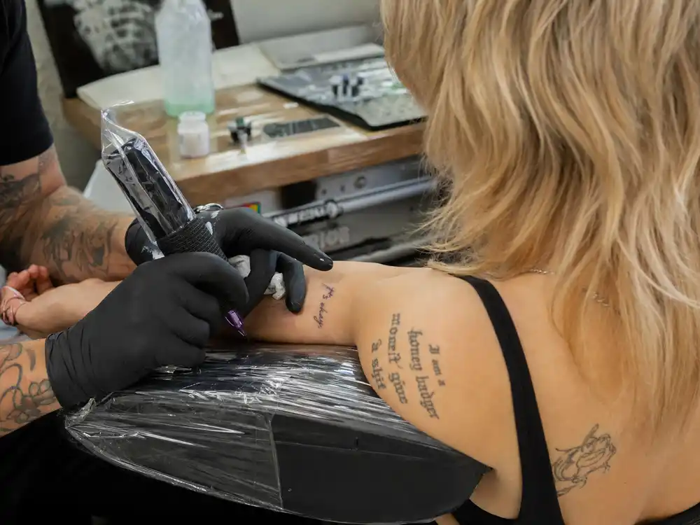 Miley Cyrus tattoos and their meanings — tattoo artist inking forearm during professional studio session with existing script tattoos visible