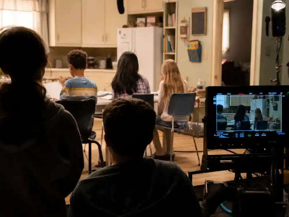 Modern Family cast net worth 2026 ranked child stars — young actor silhouettes from behind rehearsing on suburban kitchen TV set with director monitor