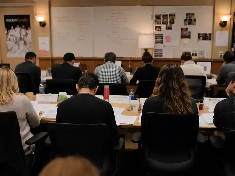 Modern Family cast net worth 2026 ranked top earners — actor silhouettes from behind at sitcom table read with scripts and coffee cups in production office