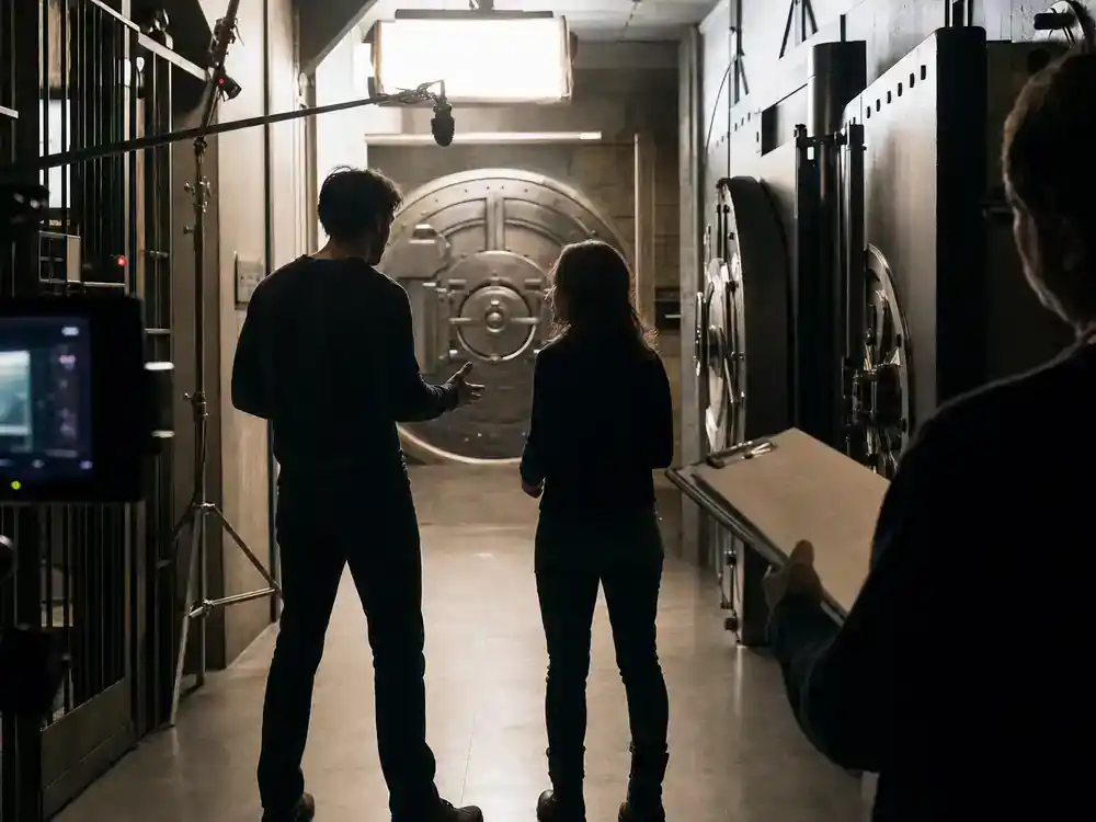 Money Heist cast and characters guide Berlin — two actor silhouettes from behind rehearsing tense dialogue inside mock bank vault corridor on TV drama set
