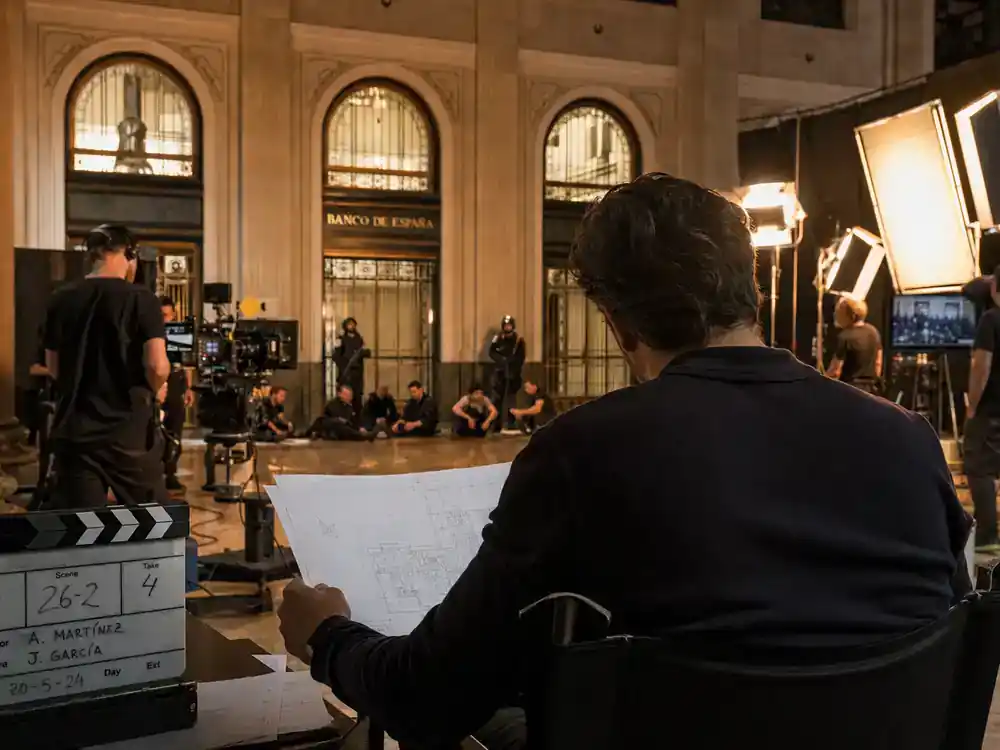 Money Heist cast and characters guide The Professor — lead actor silhouette from behind on director chair reviewing heist blueprint on Spanish bank TV set