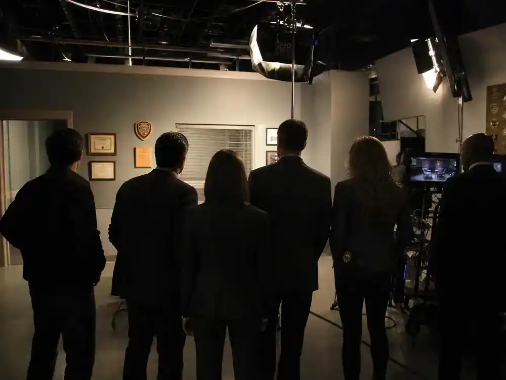 NCIS franchise 2026 cast and spinoff guide original series — ensemble TV cast silhouettes from behind on police procedural soundstage with badge props