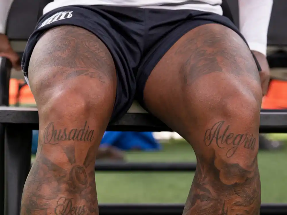 Neymar tattoos and their meanings leg knee tattoos — Ousadia and Alegria script on kneecaps training bench close-up