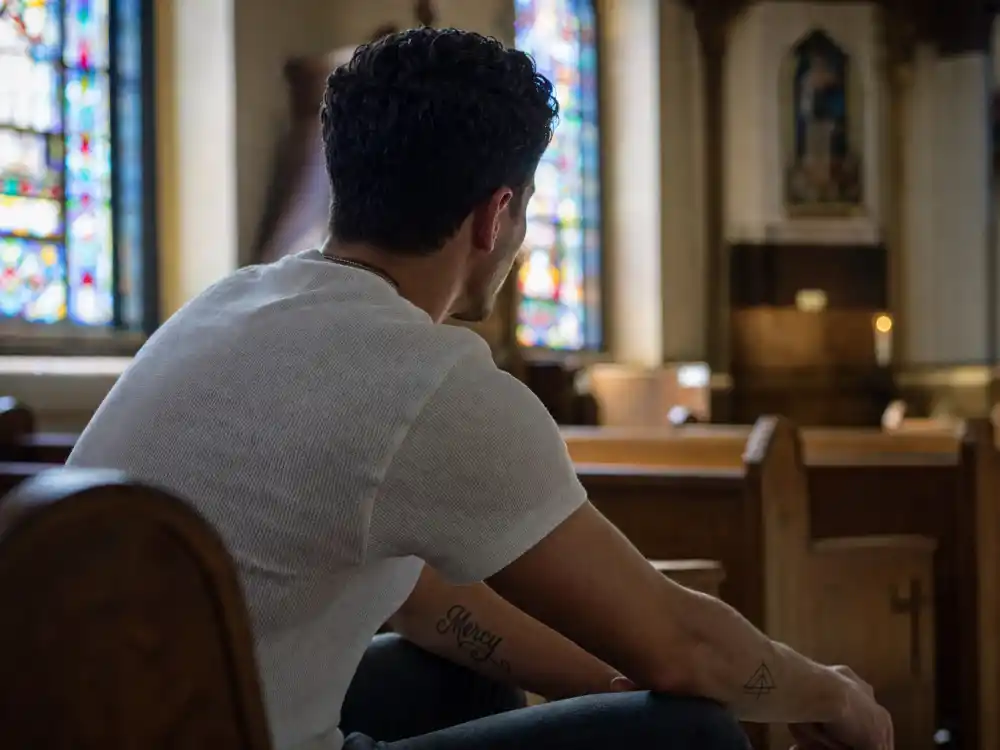 Nick Jonas tattoos and their meanings — faith and family forearm ink collection visible on church pew from behind