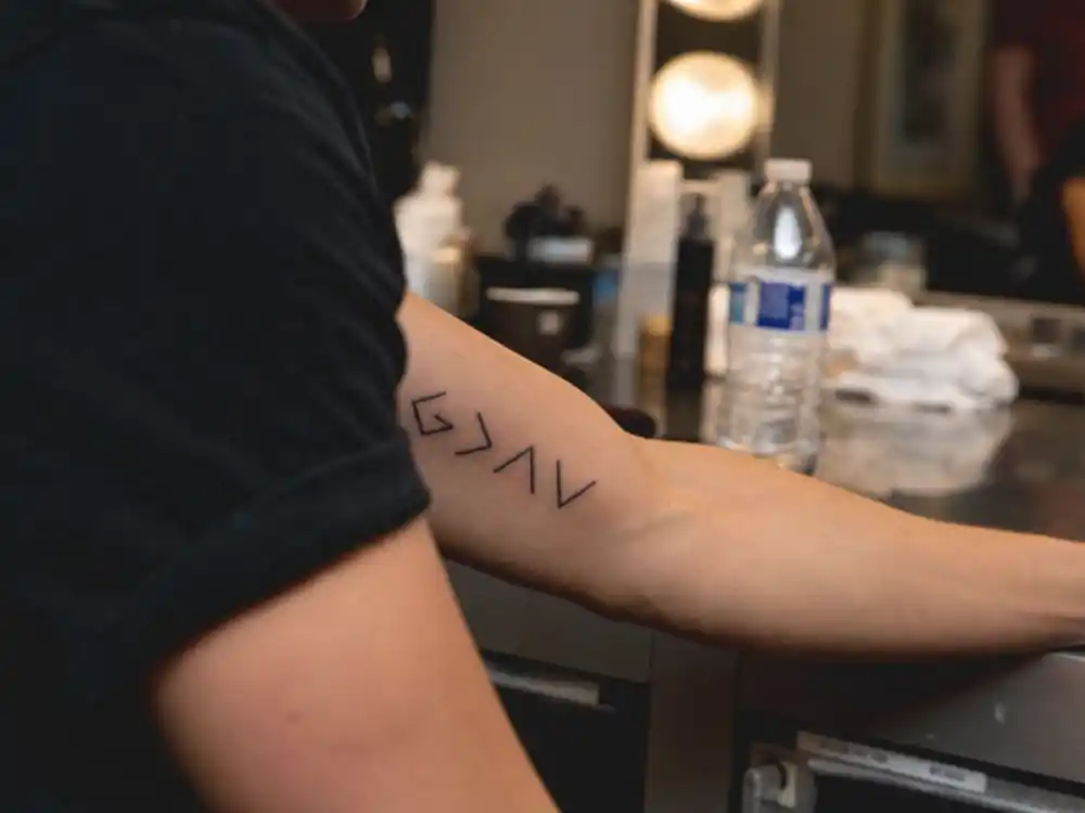 Nick Jonas tattoos and their meanings — God is greater than highs and lows geometric symbol tattoo on inner left forearm backstage