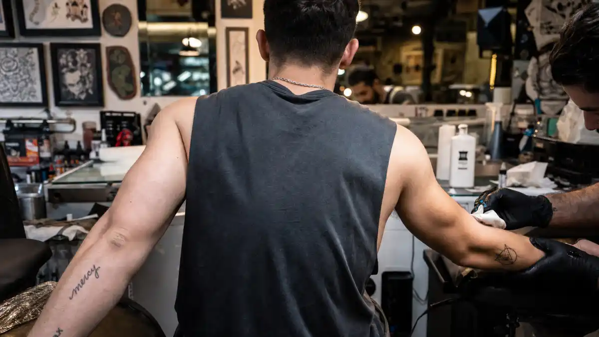 Nick Jonas tattoos and their meanings — pop musician seen from behind in tattoo studio with mercy script and geometric forearm ink visible