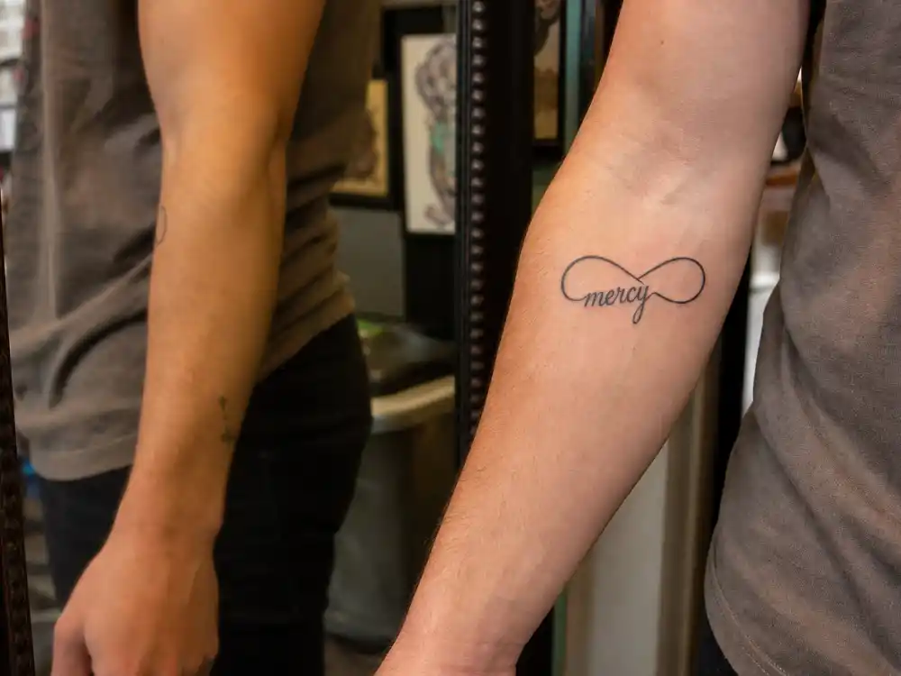 Nick Jonas tattoos and their meanings — mercy script with infinity symbol on right forearm close-up in tattoo studio mirror