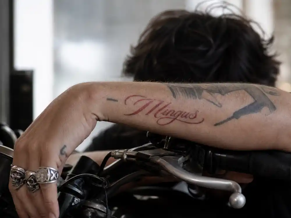 Norman Reedus tattoos and their meanings — close-up of red Mingus script forearm tattoo on motorcycle handlebar grip