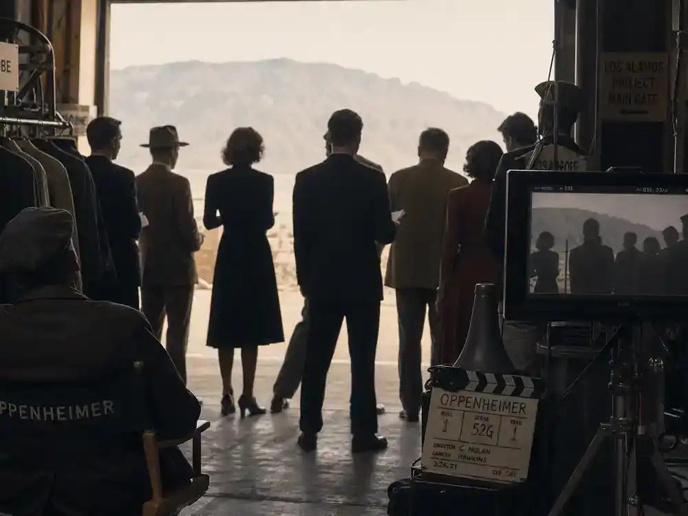 Oppenheimer movie filming locations guide ensemble cast — period-drama cast silhouettes from behind rehearsing on 1940s film set with wardrobe racks and director monitor