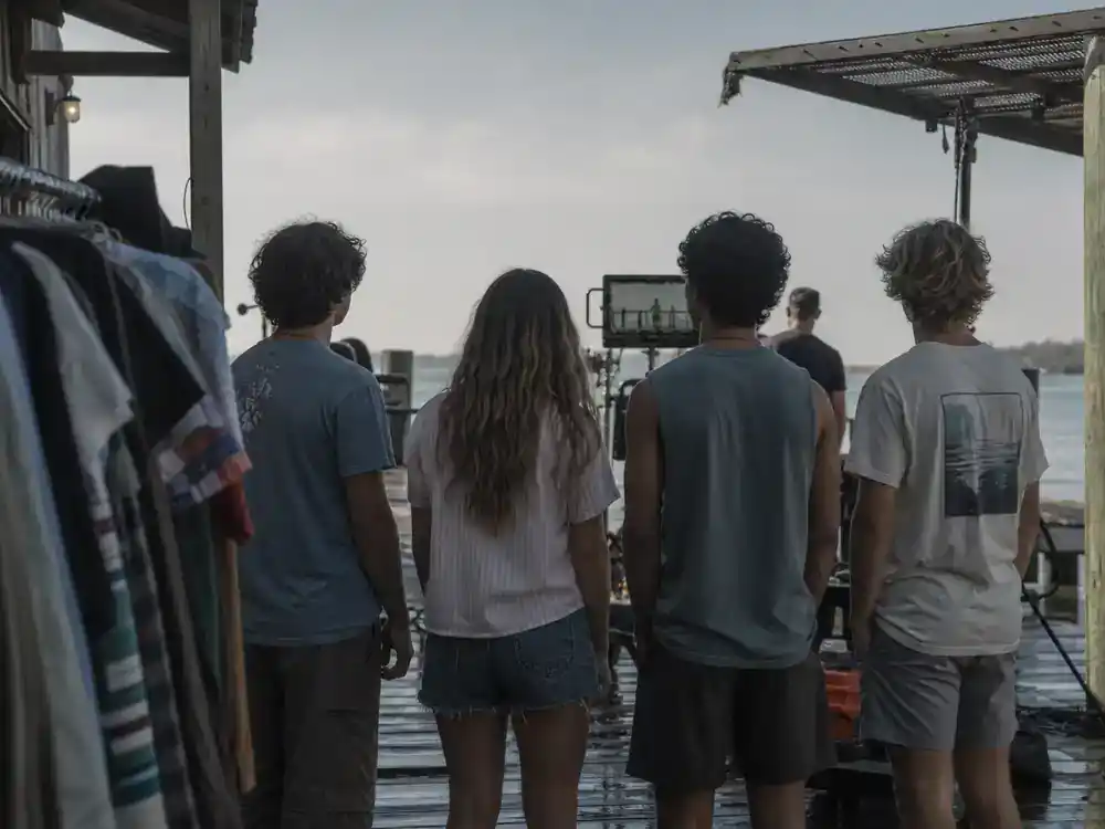 Outer Banks season 5 cast and filming guide Pogue cast — teen drama cast silhouettes from behind rehearsing on coastal dock film set with wardrobe racks