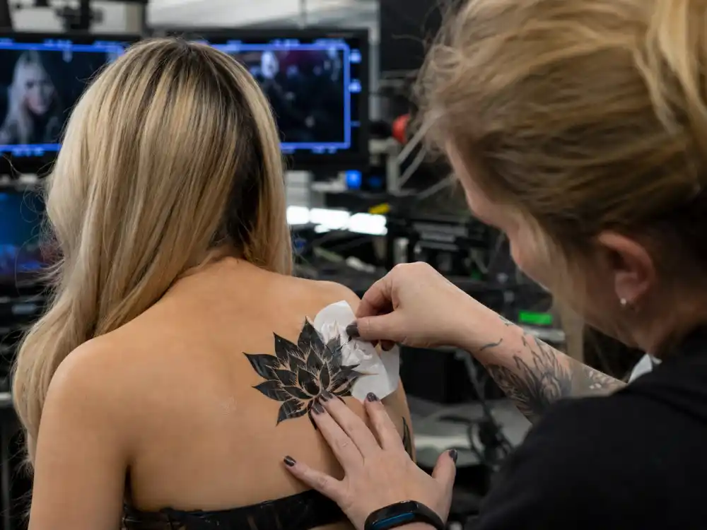 Peyton List tattoos and their meanings Black Lotus — makeup artist applying temporary black lotus tattoo transfer on film set upper back