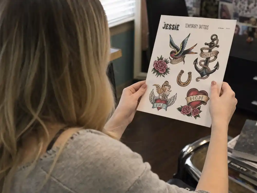 Peyton List tattoos and their meanings Jessie episode — blonde actress from behind holding temporary tattoo sheet with bare uninked forearms
