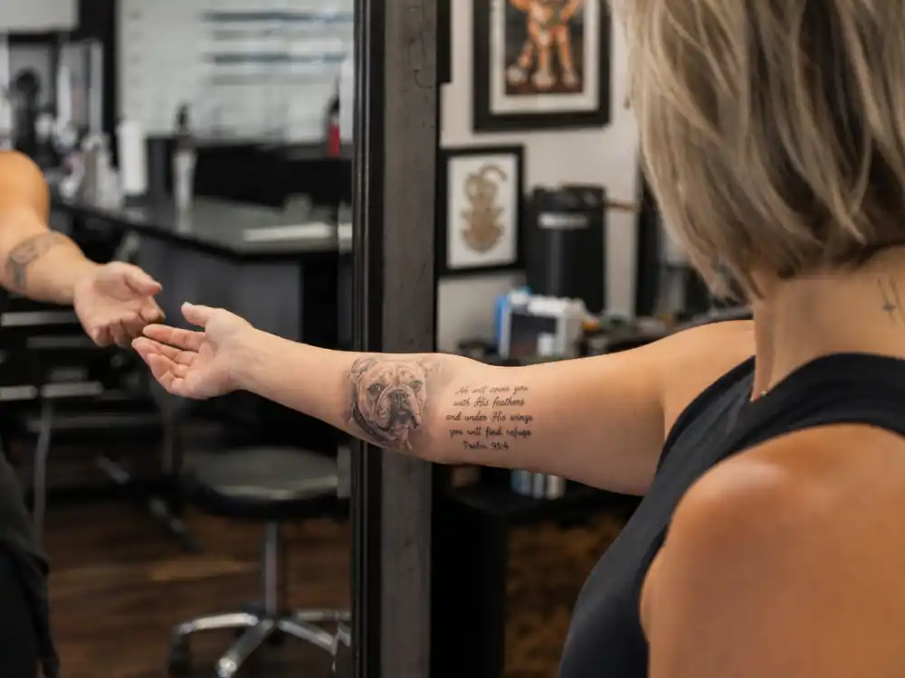 Pink tattoos and their meanings — Elvis bulldog memorial portrait and Bible verse forearm tattoo in studio mirror