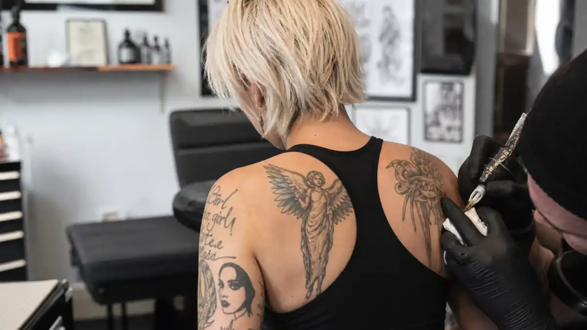 Pink tattoos and their meanings — pop star seen from behind in tattoo studio with guardian angel shoulder ink and arm tattoos visible