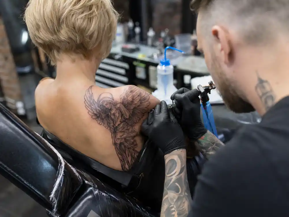 Pink tattoos and their meanings — tattoo artist inking guardian angel shoulder tattoo during professional studio session