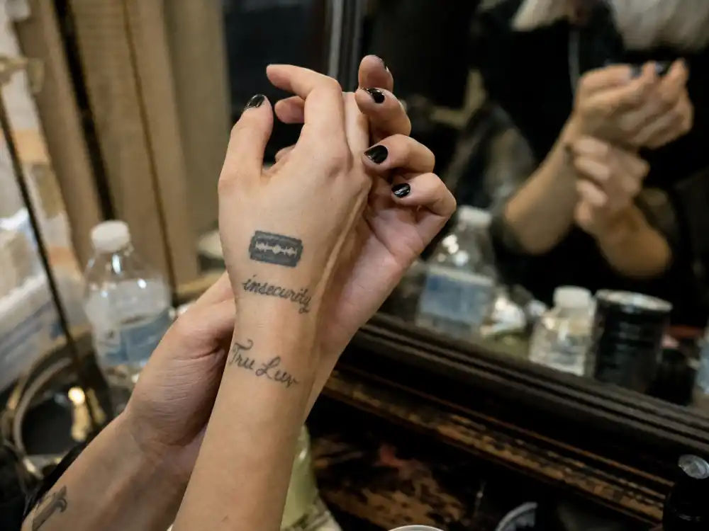Pink tattoos and their meanings — razor blade and insecurity wrist tattoos with Tru Luv script in dressing room light