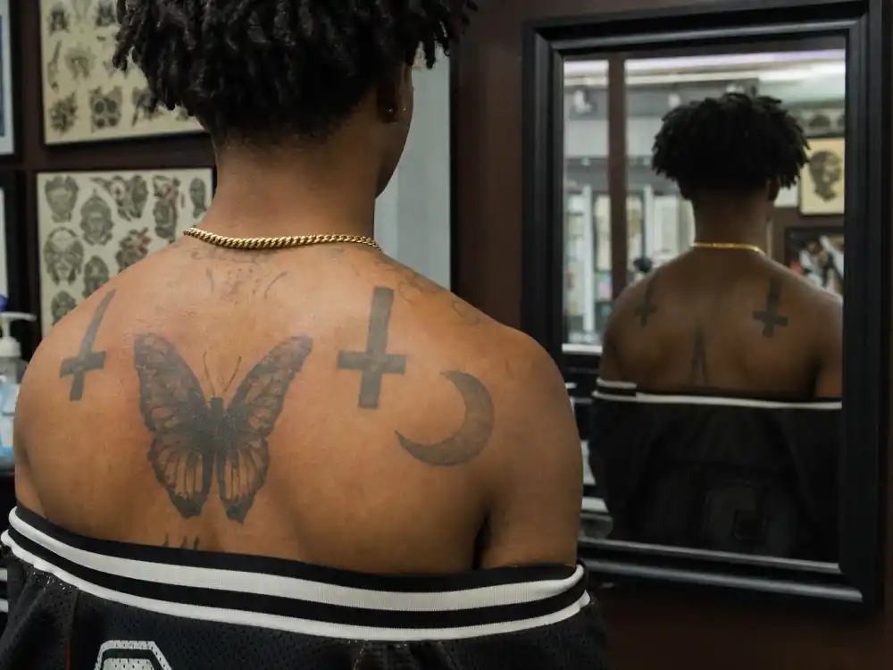 Playboi Carti tattoos and their meanings — chest butterfly tattoo with upside-down cross shoulder ink in tattoo studio mirror