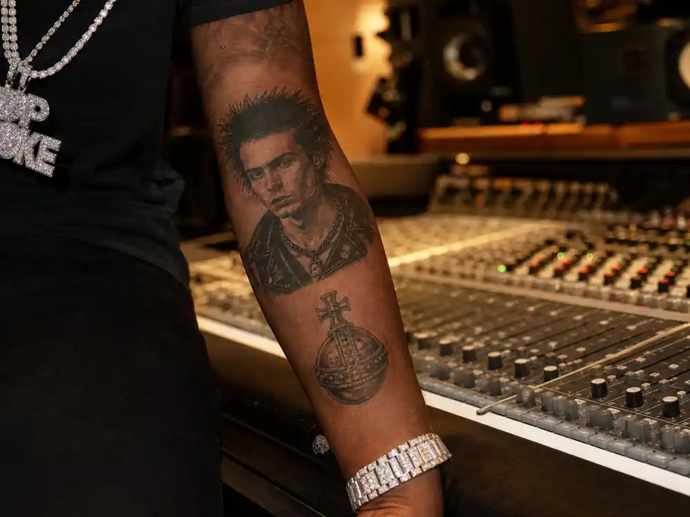 Playboi Carti tattoos and their meanings — Sid Vicious punk portrait forearm tattoo and globus cruciger ink in recording studio
