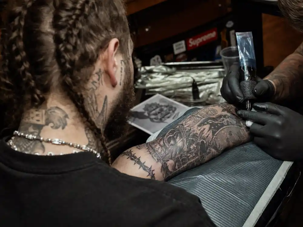 Post Malone tattoo collection guide arm sleeve — over-shoulder view of braided musician receiving forearm tattoo in studio session with artist gloved hands
