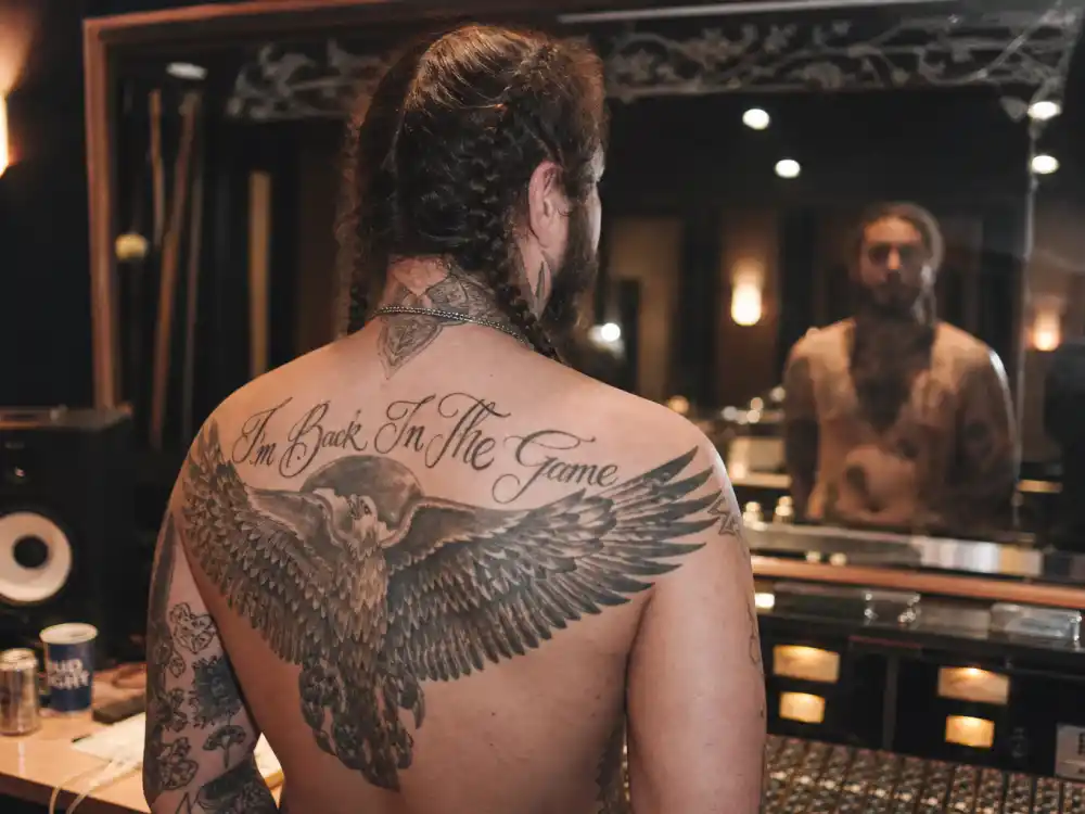Post Malone tattoo collection guide back piece — musician with braids from behind showing eagle wing shoulder tattoo in recording studio mirror
