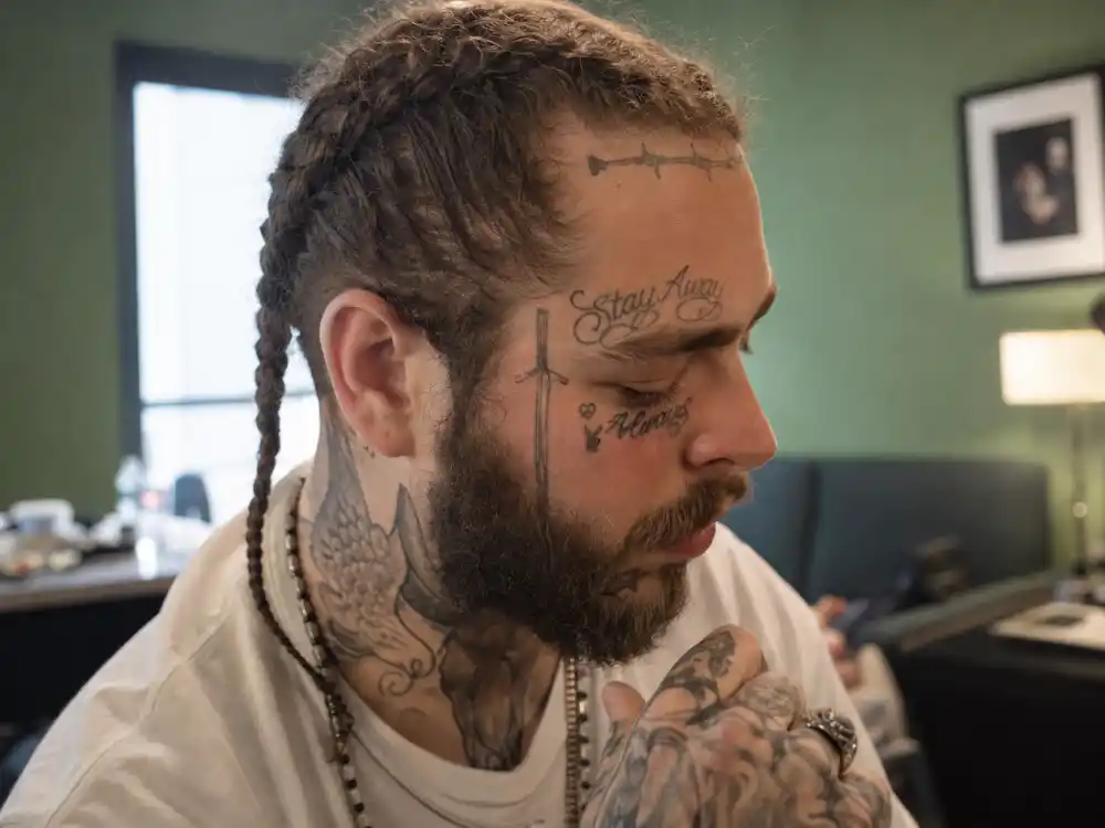 Post Malone tattoo collection guide face ink — tattooed hands and neck script tattoos of musician with braids in green room, side angle no identifiable face