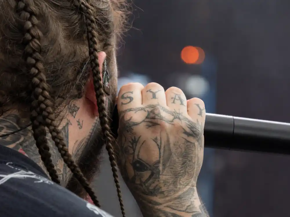 Post Malone tattoo collection guide hand ink — close-up of tattooed fingers holding microphone with knuckle letter tattoos and braids visible from behind
