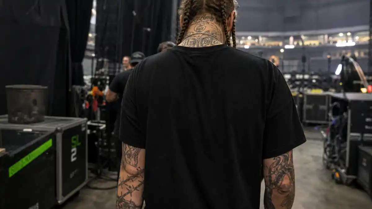 Post Malone tattoo collection guide — musician with braided hair from behind showing barbed wire forearm tattoos and neck ink backstage at arena