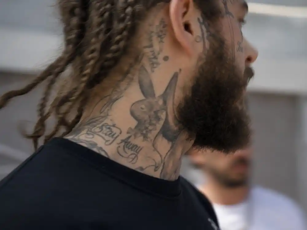 Post Malone tattoo collection guide neck tattoos — motion-blurred side profile of braided musician showing heavy facial and neck script ink outdoors