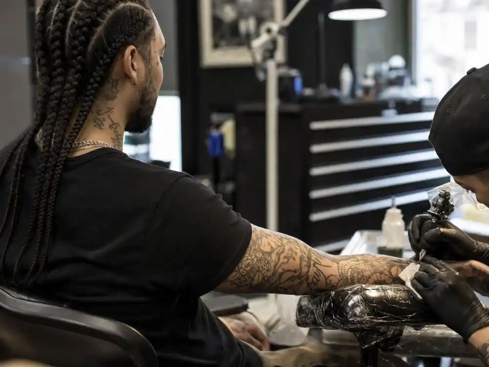 Post Malone tattoos and their meanings arm sleeve — over-shoulder view of braided musician receiving forearm tattoo in studio session with artist gloved hands